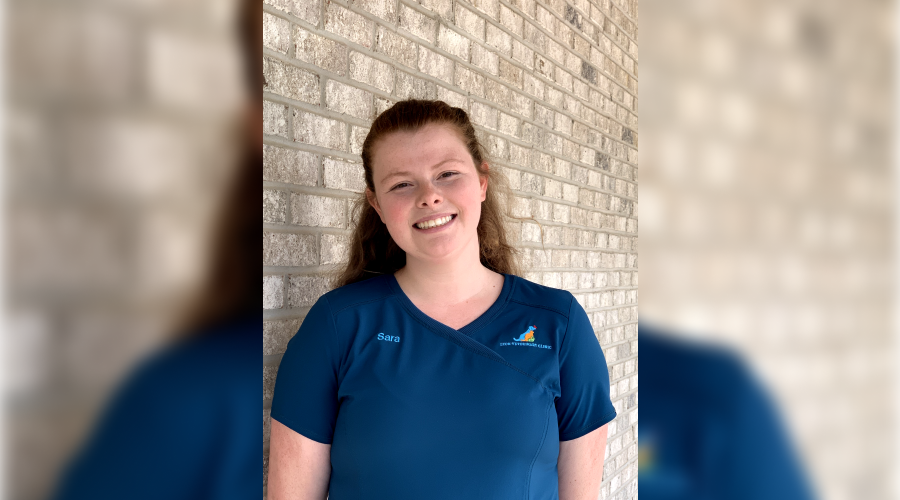 Young woman in a blue scrub top smiling in front of a brick wall.