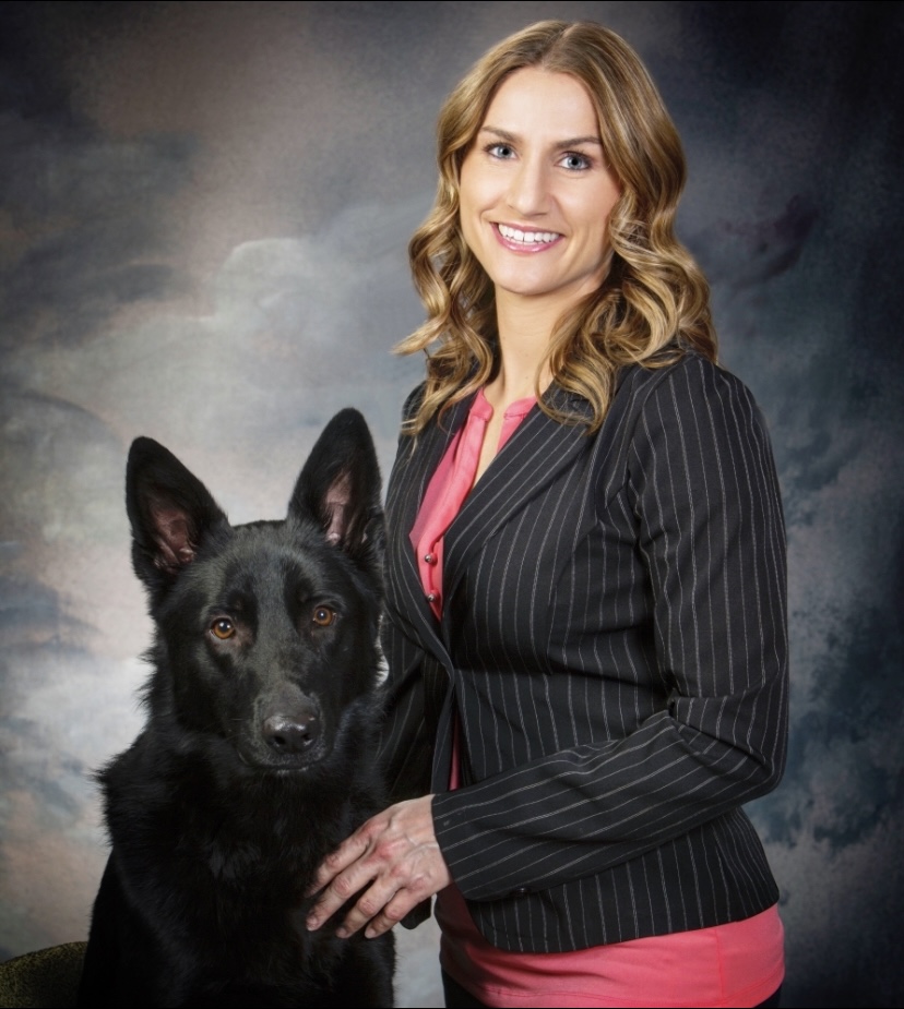 Woman in striped suit smiling with a black German Shepherd dog.