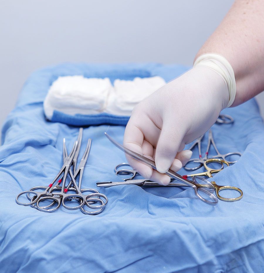 Hand in surgical glove picking up scissors from a tray of medical instruments.